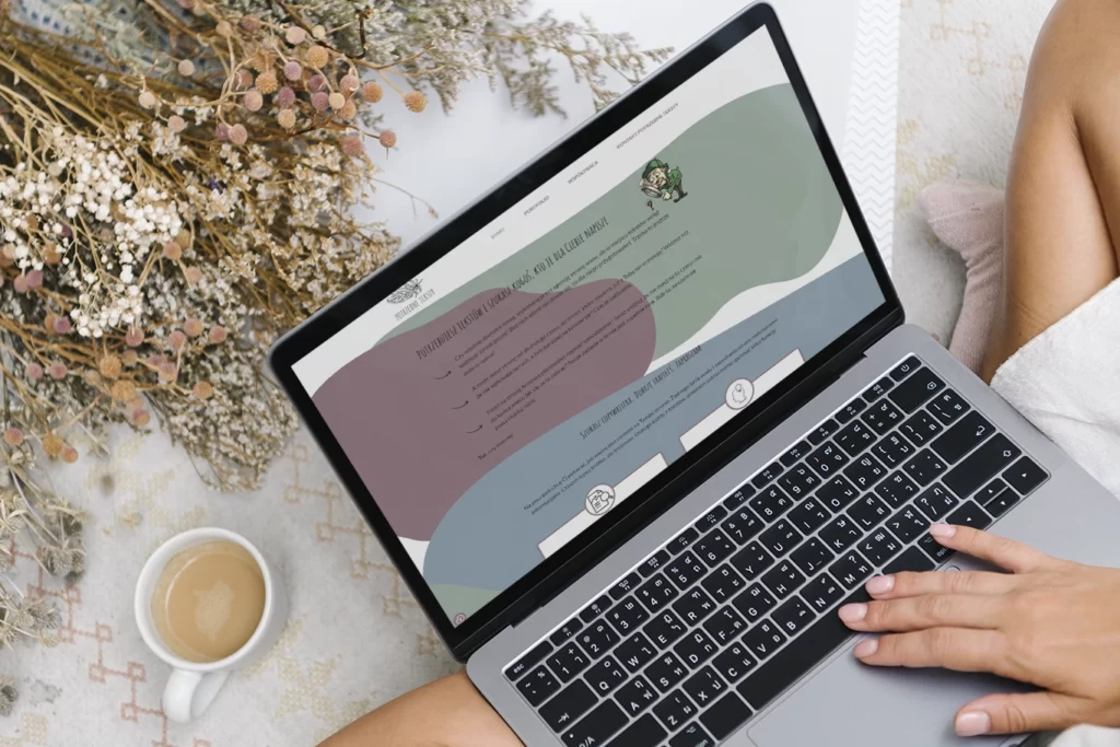Kobieta trzymająca na kolanach laptopa i przeglądająca stronę internetową https://potrzebneteksty.pl obok filiżanka z kawą i bukiet suszonych kwiatów