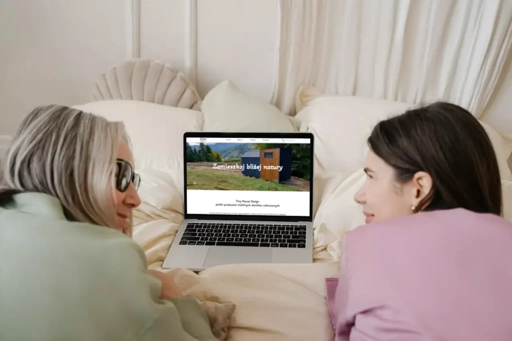 Dwie kobiety leżące na łóżku i przeglądające ofertę strony internetowej https://tinyhousedesign.pl